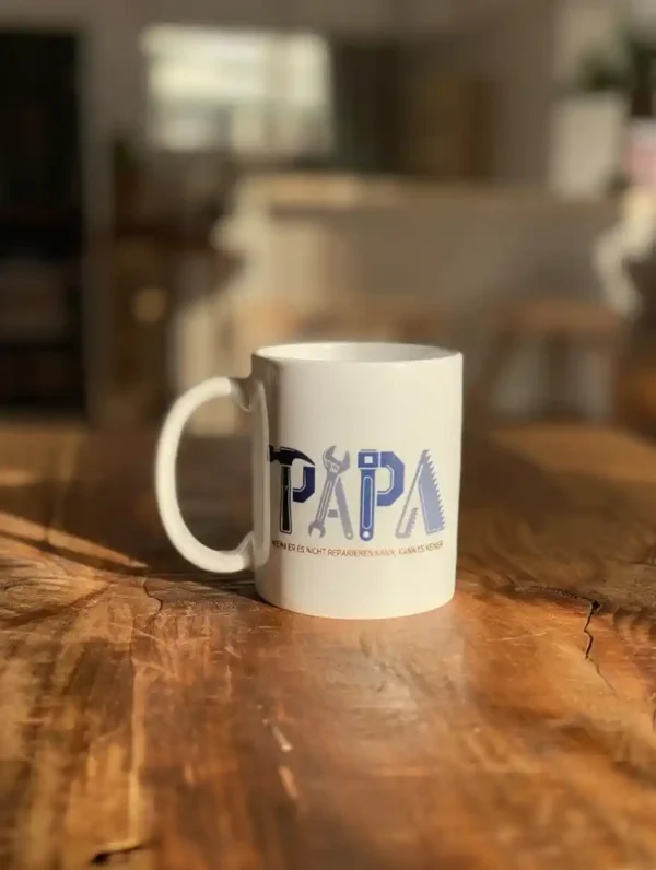 Weiße Keramiktasse mit dem Spruch 'Papa – wenn er es nicht reparieren kann, dann kanns keiner' und einer Grafik von zwei gekreuzten Schraubenschlüsseln auf Holztisch.